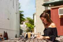 woman writing in a journal and iced coffee and cappuccino on a wooden table outdoors 