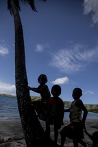 Silhouette of children leaning on a palm tree by the ocean.