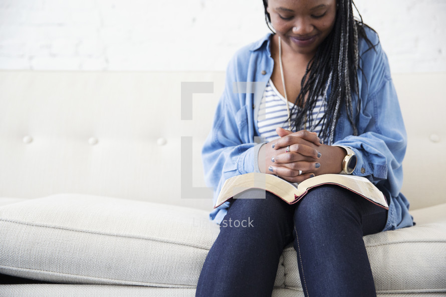 a young woman sitting on a couch reading a Bible and praying 