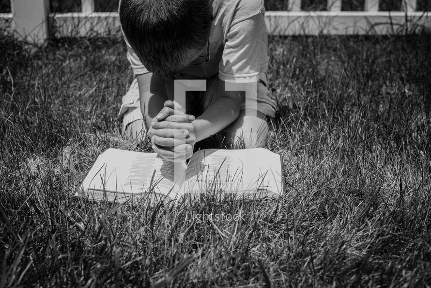 a boy kneeling in the grass reading a Bible God's Word  and praying 