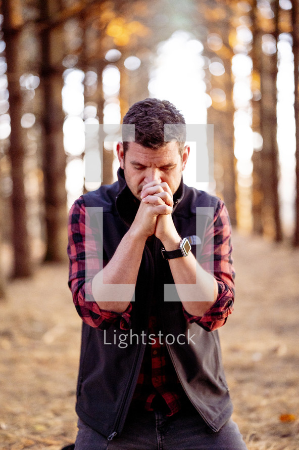 a man kneeling in a forest with praying hands 