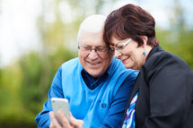 elderly couple sitting on a park bench with a cellphone 