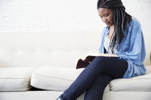 A young woman sitting on a couch reading a Bible.