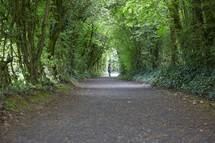 a woman walking on a gravel road under a canopy of trees