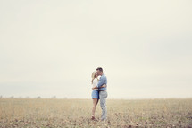 A man and woman embrace while standing in a field.
