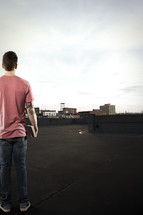 Man holding a Bible standing on a rooftop, looking at skyline of buildings.