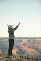 a young man standing on the edge of a cliff with hands raised in praise 