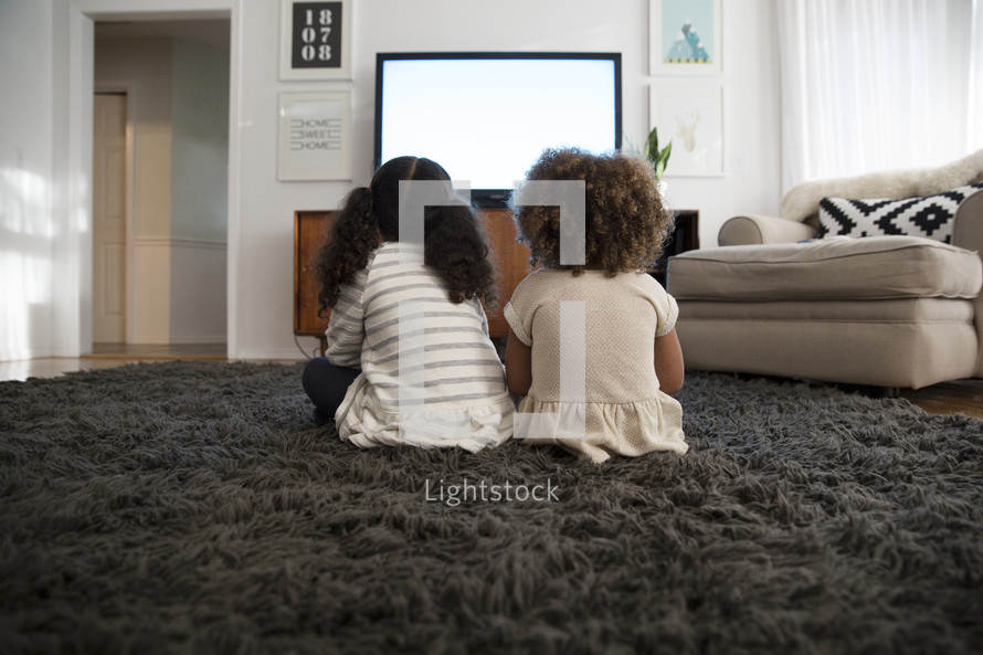 girls watching tv, sitting on a rug in a living room 