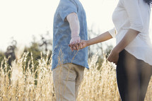 a couple walking holding hands outdoors through tall grasses 