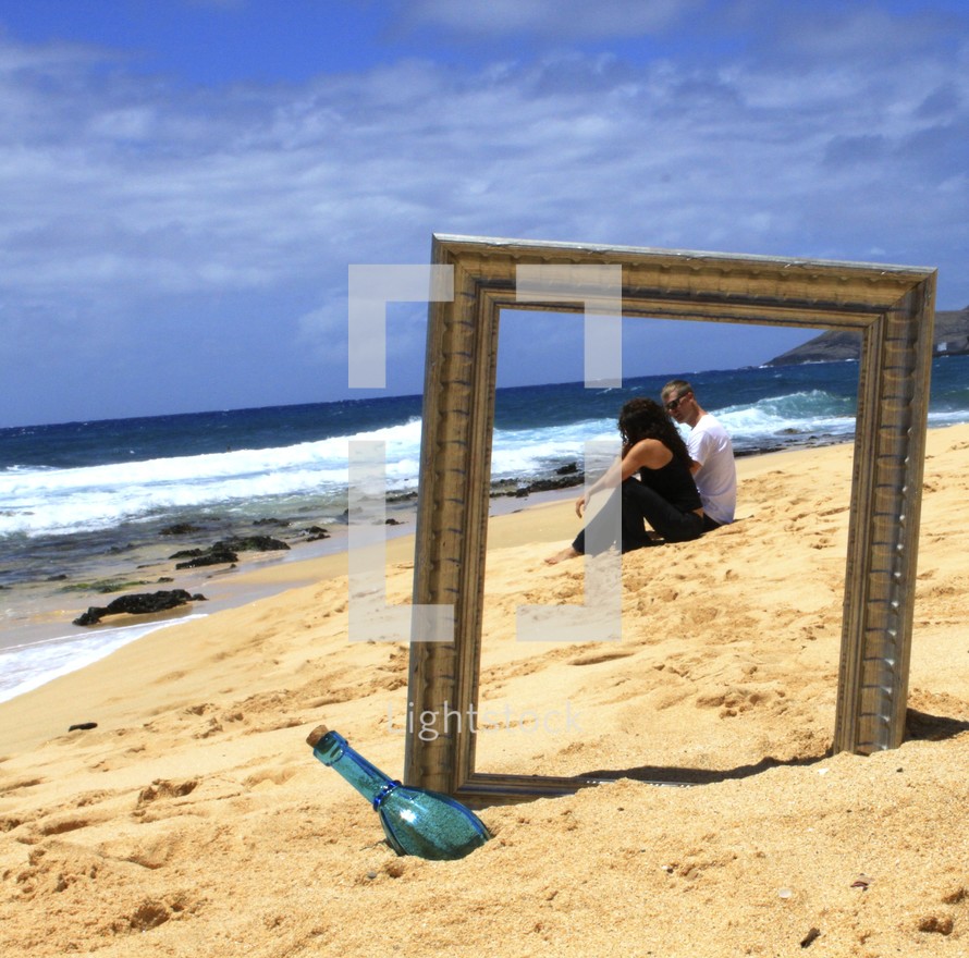 Couple sitting on the beach as seen through a frame near a glass bottle.