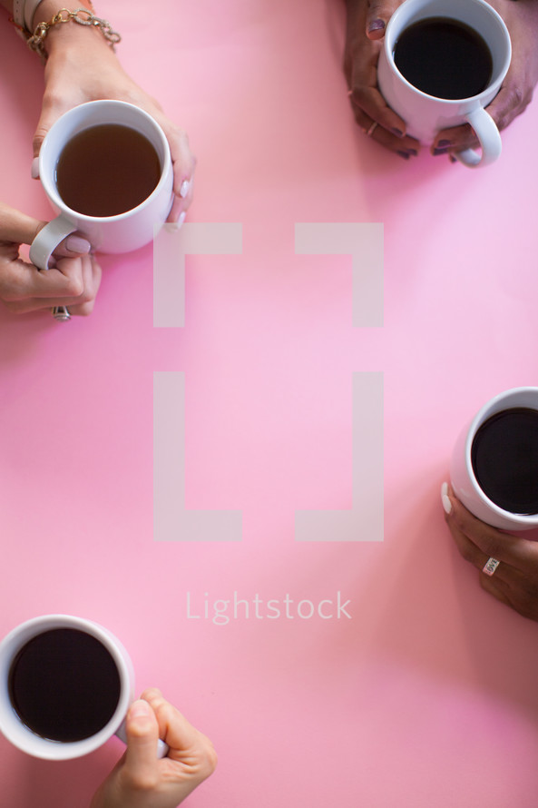 The hands of four women holding coffee cups on a pink table.