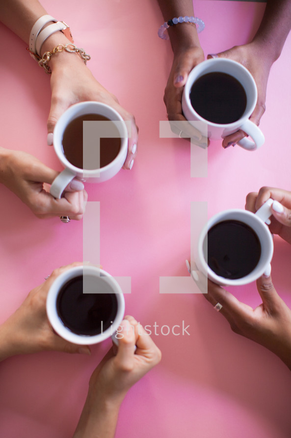 The hands of four women holding coffee cups at a pink table.