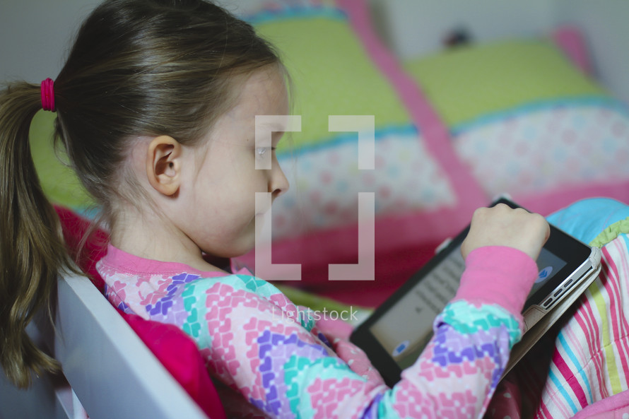 Girl in pajamas reading the Bible on an iPad and smiling. She's reading before bedtime.  Child doing devotions on a tablet. Reading a children's Bible on a tablet.