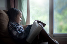 a toddler girl sitting in a window reading a Bible 