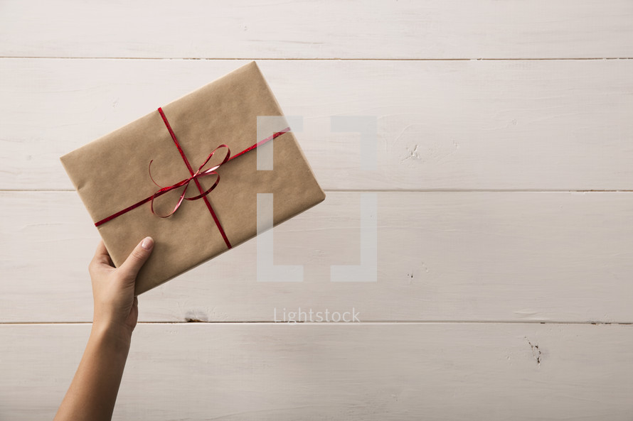 A gift wrapped in brown paper with a red ribbon being placed on a table.