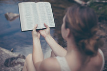 woman sitting on a rock reading a Bible by a river 
