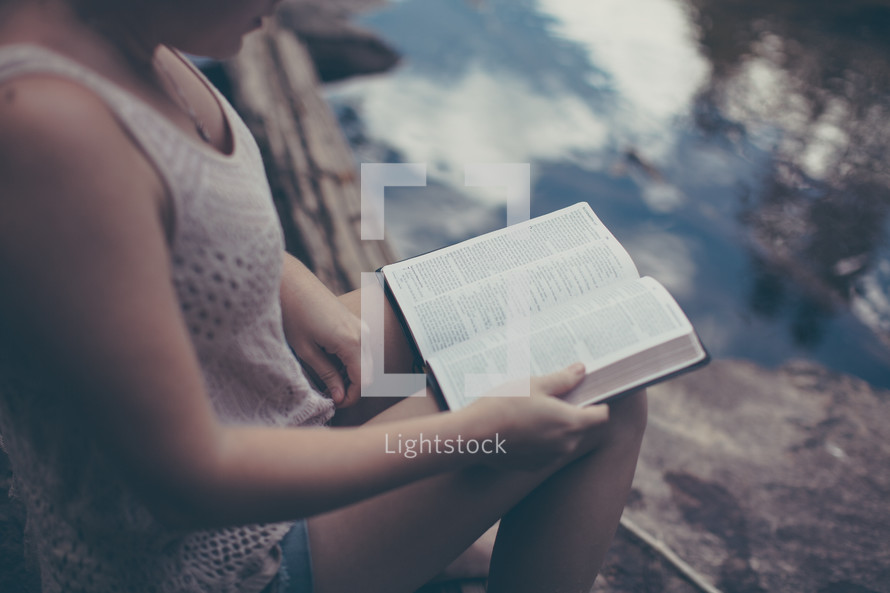 woman sitting on a rock reading a Bible by a river 
