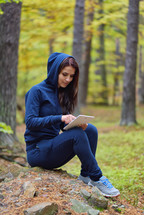 a woman sitting in a forest looking at a tablet screen 