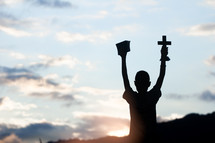 silhouette of a boy holding a cross and Bible with hands raised 