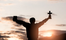 a silhouette of a boy holding a cross and Bible at sunrise 