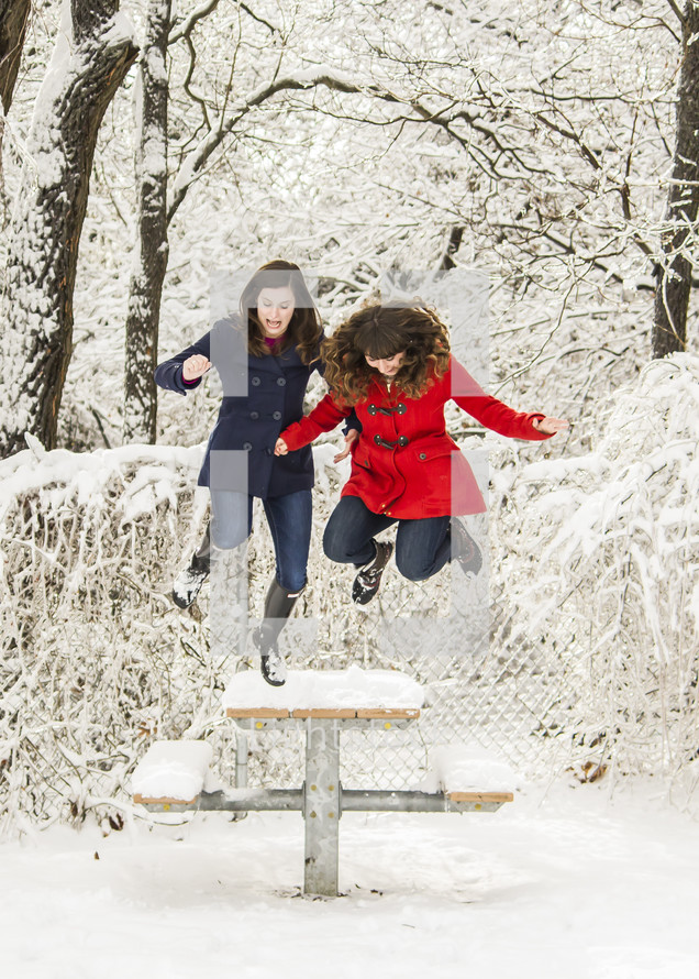 Snowy trees with two girls jumping off a picnic bench covered with snow .
