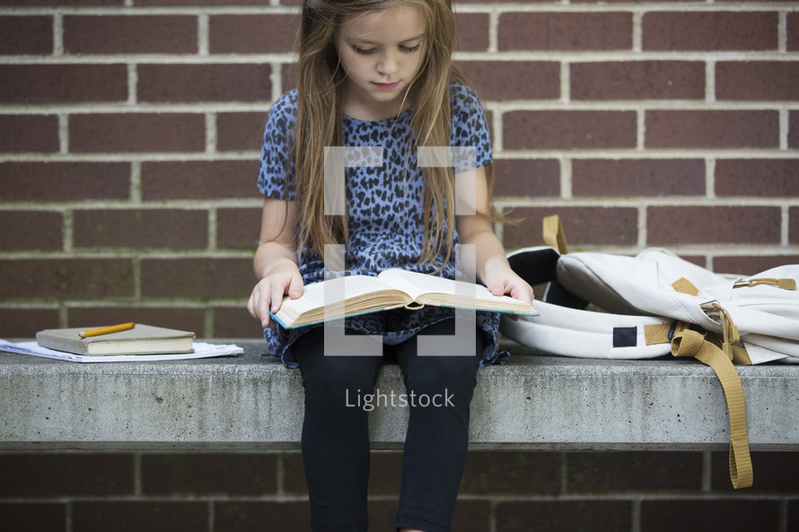 a child sitting on a bench reading a book while waiting for school to start 