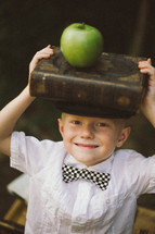 a boy child with a stack of books on his head and an apple 