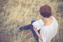 Woman sitting outside in a field of grass reading the Bible.