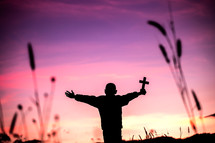 boy holding up a cross with sunlight at sunset in the background