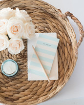 A pencil and notecard, candle, and white flowers on a rattan basket.
