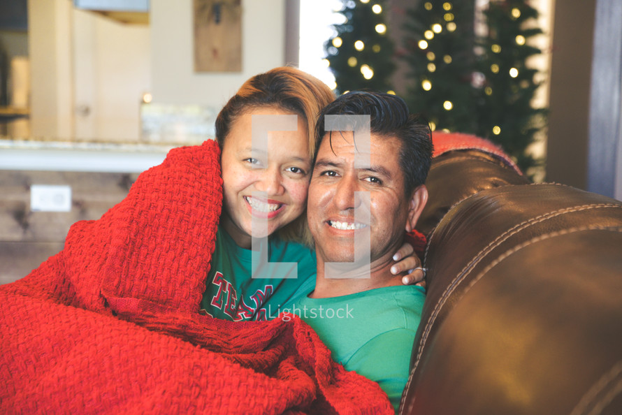 a couple snuggling under a blanket sitting on a couch at Christmas 