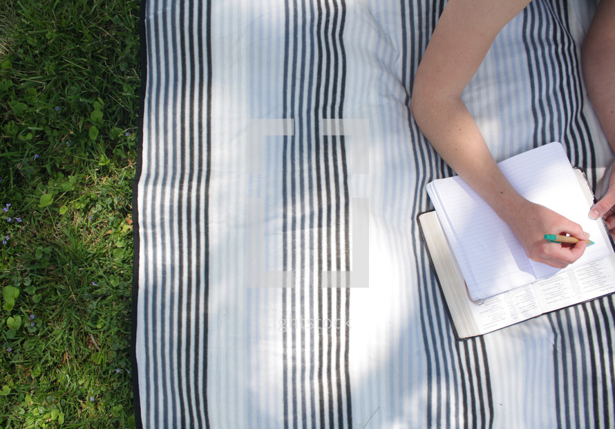 a girl lying on a blanket in the grass reading a Bible and writing in a journal 