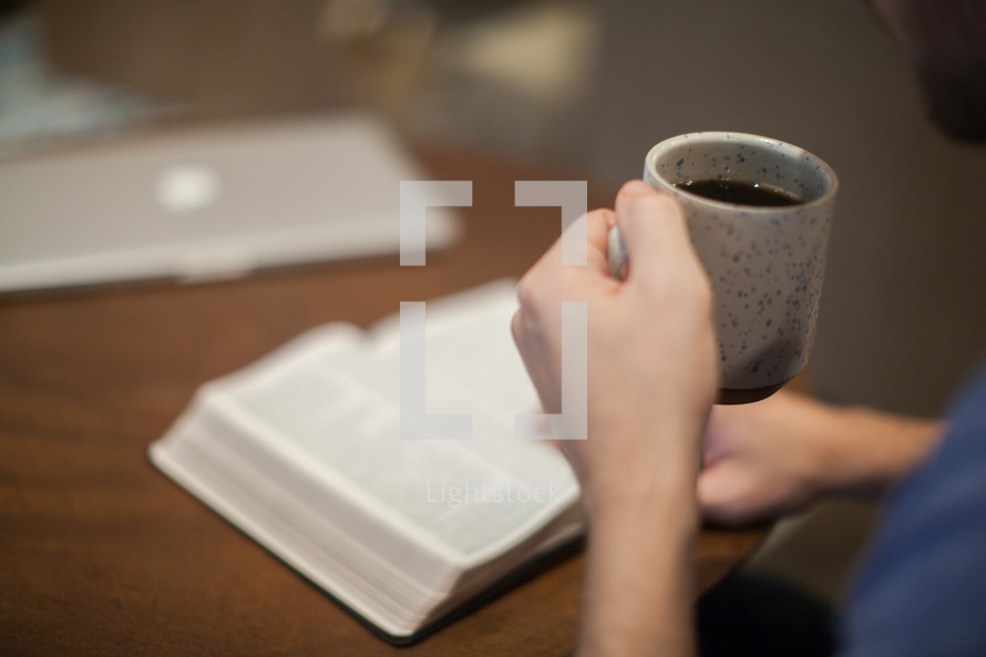 A young man sitting at a table reading the Bible holding a cup of coffee