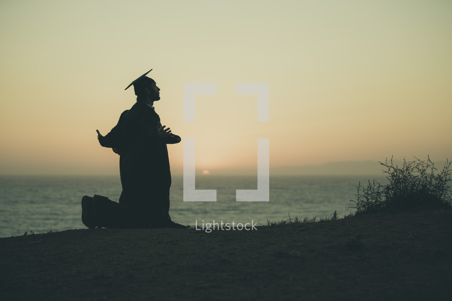 silhouette of a graduate on a beach kneeling in prayer