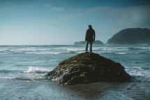 a man standing on a rock on a beach looking out at the ocean 