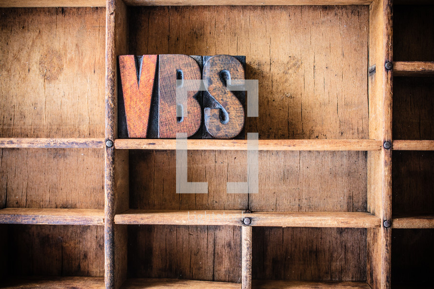Wooden letters spelling "VBS" on a wooden bookshelf.