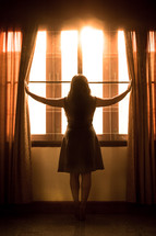 a woman pushing open curtains to look out a window 