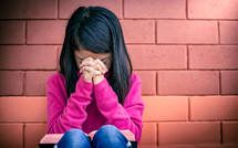 a girl child praying in front of a brick wall 