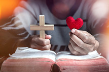 a man holding a cross and red felt heart over the pages of a Bible 