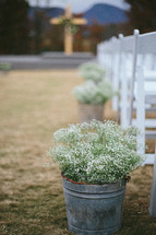 Buckets of baby's breath flowers at then end of rows of chairs, with an aisle leading to a cross.