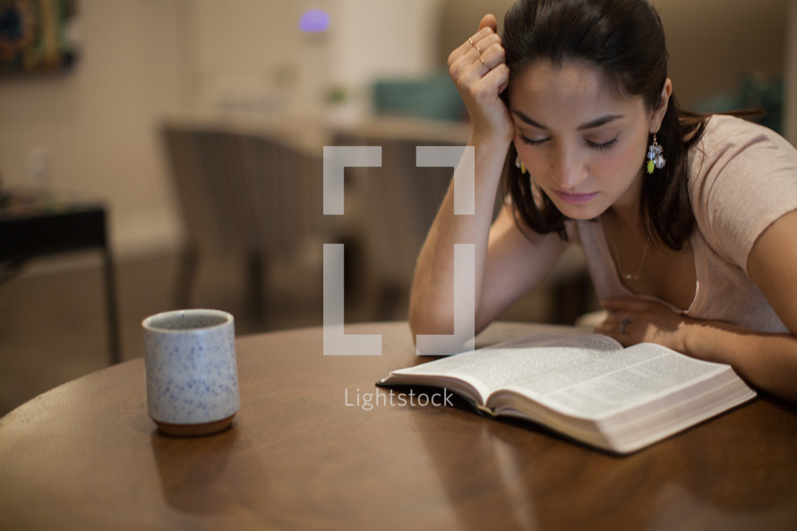A young woman sitting at a table reading the Bible looking tired