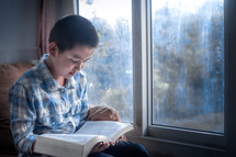 a boy reading a Bible sitting in a window 