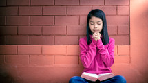 a girl reading a Bible and praying in front of a brick wall 