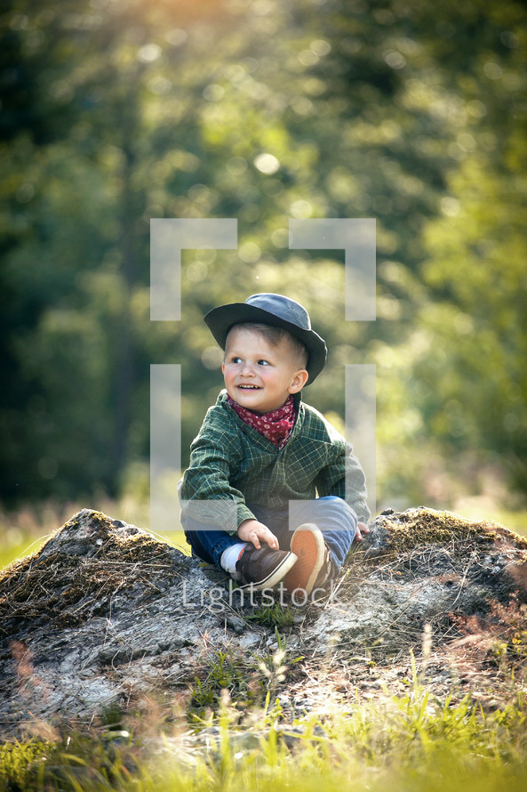 Cute Little Boy with Hat and Checkered Shirt Sitting in Summer Forest