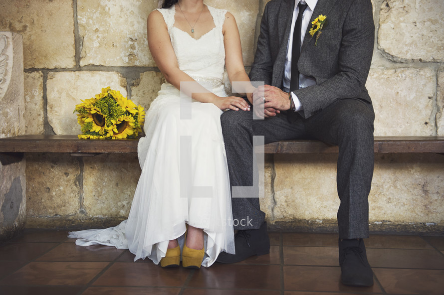 Bride with sunflower bouquet sitting on wooden bench holding hands with groom in chapel.