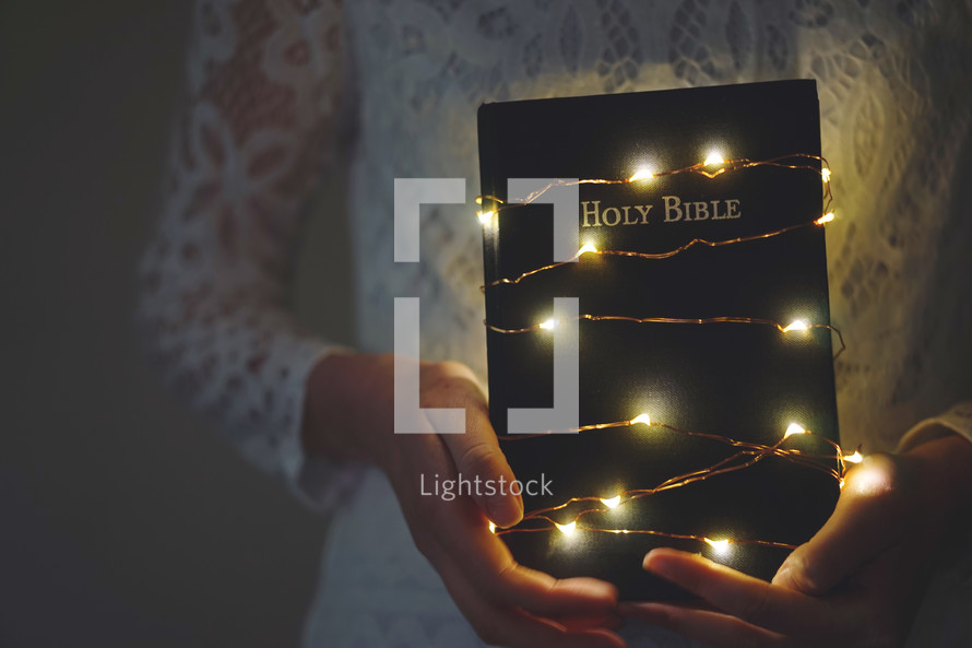 a woman holding a Bible wrapped in fairy lights 