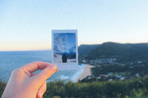 woman holding up a polaroid picture near a coastline 