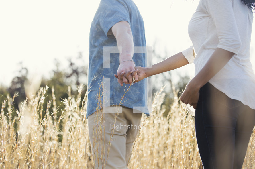 a couple walking holding hands outdoors through tall grasses 