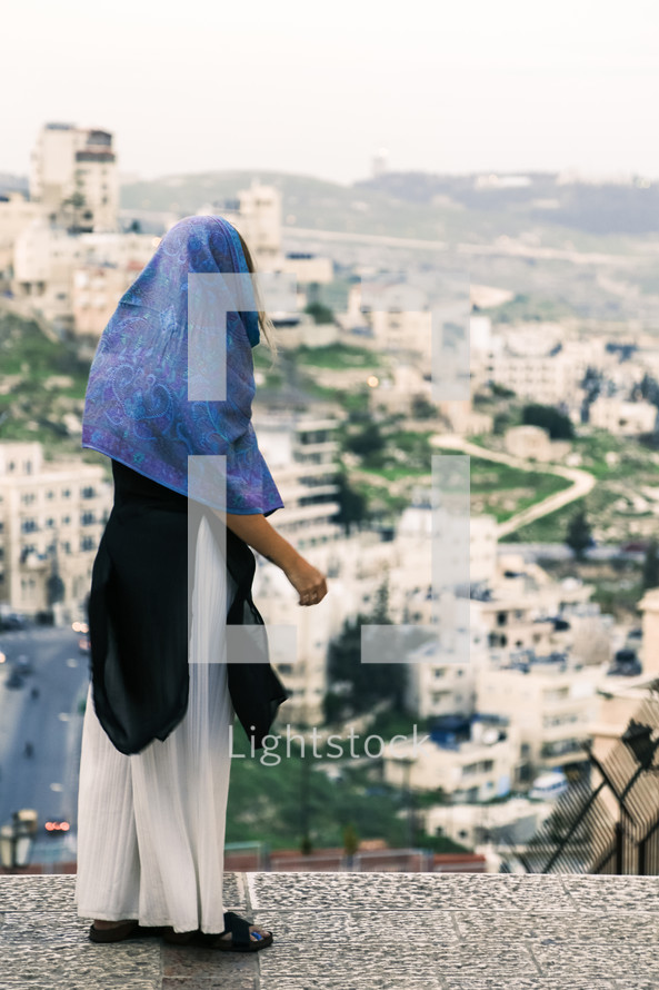 a woman with a scarf over her head looking out at town below 
