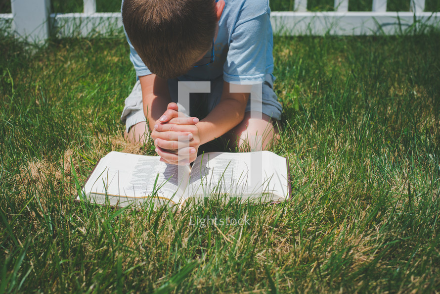 a boy kneeling in the grass reading a Bible and praying 